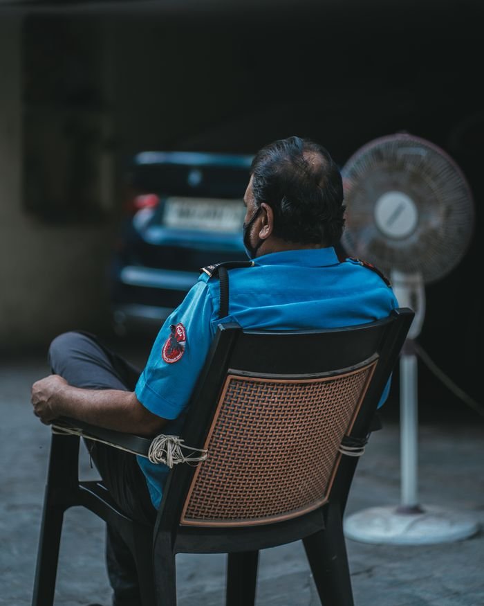 Back view of a security guard sitting outdoors, wearing a blue uniform and black mask, near a parked car.